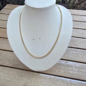 May include: A gold-colored chain necklace displayed on a white bust. The necklace has a flat, herringbone design. The bust is set on a wooden table, with a natural outdoor background.