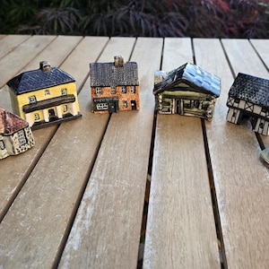May include: A selection of miniature ceramic houses, each with unique architectural details. The houses are painted in a variety of colours, including yellow, brown, and black, and are displayed on a wooden surface. The houses feature different roof styles and window designs.
