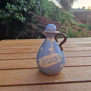 Puede incluir: Una botella de vinagre de cerámica azul y blanca con lunares blancos y la palabra "Vinagre" escrita en blanco sobre una pancarta amarilla. La botella tiene un asa marrón y una tapa marrón.