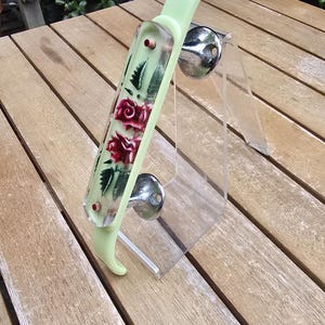 Könnte beinhalten: Ein hellgrüner Schuhlöffel mit einem rechteckigen, transparenten Einsatz, der mit roten Rosen und grünen Blättern verziert ist. Der Griff hat zwei kleine rote Akzente und ist auf einem klaren Acrylständer mit zwei runden Chromstützen montiert.