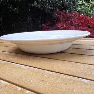 May include: A white, shallow, ceramic serving bowl with a wide rim. The bowl is set on a wooden table with a natural finish. The background features lush greenery and red foliage, suggesting an outdoor setting.