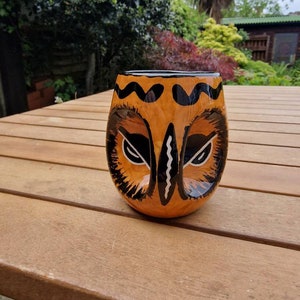 Könnte beinhalten: Eine Keramiktasse in Form eines Eulenkopfes. Die Tasse ist orange mit schwarzen und weißen Details. Die Augen der Eule sind schwarz mit weißen Pupillen. Die Tasse steht auf einem Holztisch.