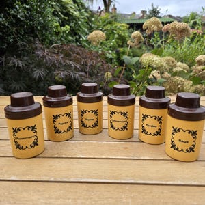May include: Set of seven brown and yellow spice jars with black labels and different spice names. The jars are arranged in a row on a wooden surface.