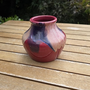 May include: A small ceramic vase with a rounded body and a wide opening. The surface features a glossy glaze with a colour palette of deep red, dark purple, and creamy beige, showing a distinct drip pattern where the colours blend together.