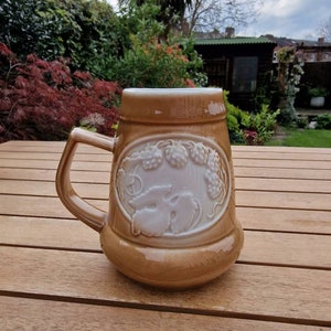 May include: A brown ceramic beer stein with a white embossed design of grapes and leaves. The stein has a handle and is sitting on a wooden table.