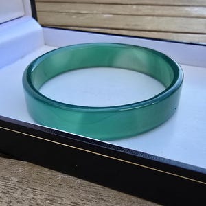 Könnte beinhalten: Ein glatter, durchscheinender grüner Armreif mit polierter Oberfläche. Das kreisförmige Schmuckstück wird in einer rechteckigen schwarzen Präsentationsbox mit weißem Innenfutter präsentiert und zeigt seine leuchtende Farbe und solide Form.