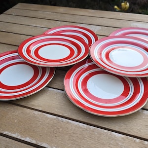 May include: A set of five round ceramic plates featuring a white center surrounded by concentric rings in bright red and thin dark blue lines. The plates have a smooth finish and are arranged in a slightly overlapping cluster on a flat surface.