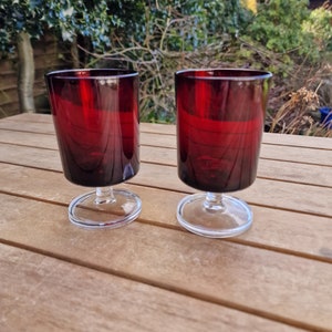 May include: Two clear glass shot glasses with a red, ruby-colored base. The glasses are on a wooden surface.