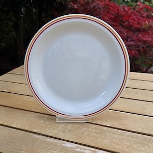 May include: A white ceramic plate with a decorative rim featuring a thin burgundy stripe between two gold stripes. The plate is displayed on a wooden table, showcasing its clean design and simple elegance. Perfect for serving meals.