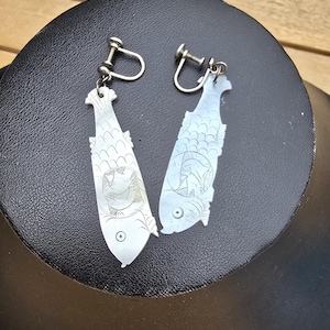 May include: A pair of silver-toned fish-shaped earrings. Each earring features a detailed fish design with etched scales, fins, and a circular eye. The earrings have a screw-back closure and are displayed on a black surface.
