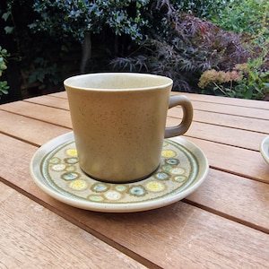 Könnte beinhalten: Eine braune Kaffeetasse aus Keramik mit einem Unterteller mit grünem und gelbem Muster. Tasse und Unterteller stehen auf einem Holztisch.