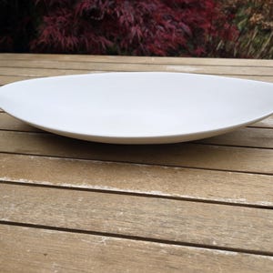 May include: A white, oval serving dish. The ceramic plate is placed on a wooden table with a natural finish. The background shows green and red foliage.