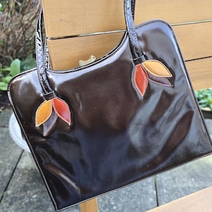 May include: A glossy, dark brown handbag with a rectangular shape and a curved top. The bag features two handles and decorative leaf-shaped accents in shades of orange, yellow, and red. The bag is sitting on a wooden bench.