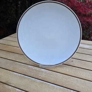 May include: A white ceramic plate with a dark brown rim. The plate is round and sits on a clear plastic stand. The plate is on a wooden table.