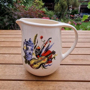 Puede incluir: Jarra de cerámica blanca con un diseño colorido de un instrumento musical, una tetera y espigas de trigo. La jarra tiene un asa y un cuerpo redondeado.