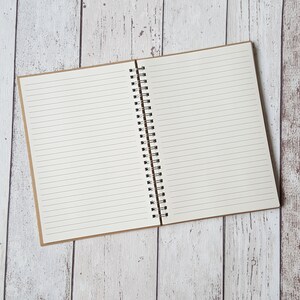 Puede incluir: Un cuaderno abierto de espiral con p&aacute;ginas rayadas. El cuaderno tiene una cubierta de papel marr&oacute;n y est&aacute; acostado sobre una superficie de madera blanca.