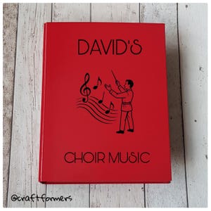 Peut inclure: Un livre rouge avec le texte "DAVID'S CHOIR MUSIC" en noir. Une illustration noire d'un chef d'orchestre avec des notes de musique est sur la couverture. Le livre est rectangulaire et semble être un livre de musique.