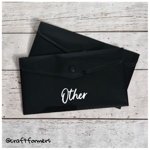 May include: Two black document envelopes with a button closure. The word "Other" is written in white script on the front of the envelope. The envelopes are on a light wood grain surface.