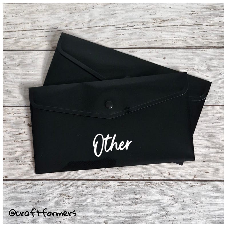 May include: Two black document envelopes with a button closure. The word "Other" is written in white script on the front of the envelope. The envelopes are on a light wood grain surface.