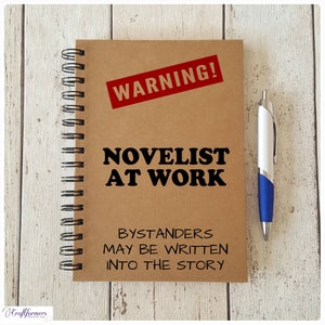 May include: A brown notebook with a spiral binding. The cover has a red warning sign that says "WARNING!" and the text "NOVELIST AT WORK BYSTANDERS MAY BE WRITTEN INTO THE STORY". A blue pen is lying next to the notebook.