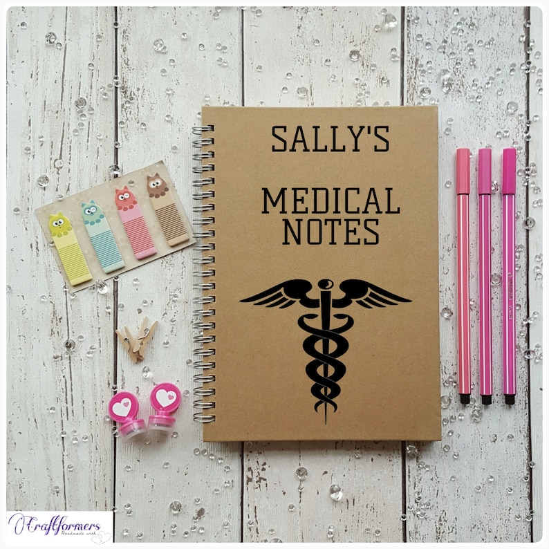 Puede incluir: Una libreta espiral marr&oacute;n con el texto "SALLY'S MEDICAL NOTES" y un s&iacute;mbolo m&eacute;dico negro. La libreta est&aacute; sobre una superficie de madera blanca con bol&iacute;grafos rosas y notas adhesivas de colores.