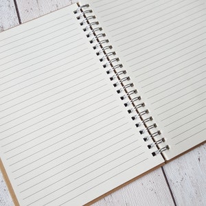 May include: A white spiral bound notebook with lined pages. The notebook is open and lying flat on a light wood surface.