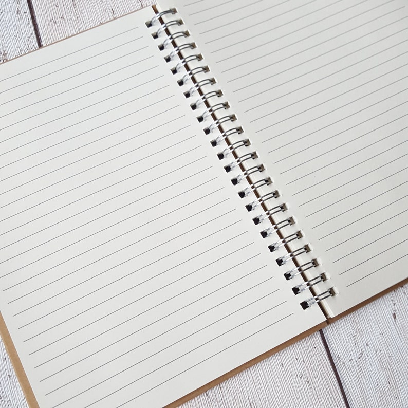 May include: A white spiral bound notebook with lined pages. The notebook is open and lying flat on a light wood surface.