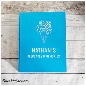 May include: A vibrant turquoise keepsake book featuring white text and a balloon design. The cover displays "NATHAN'S KEEPSAKES & MEMORIES". The balloons have star, dot, and swirl patterns. The book is rectangular and rests on a light wood surface.