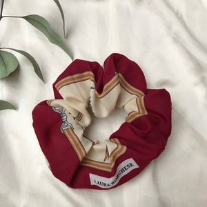 Puede incluir: Scrunchie de tela con estampado rojo y crema con el texto "LAURA BORGHESE" impreso en la parte inferior.