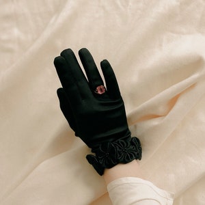 Könnte beinhalten: Ein schwarzer Satinhandschuh mit floraler Verzierung und einem rosafarbenen Ring mit einem quadratischen Stein.