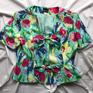 May include: A short-sleeved blouse with a floral print in shades of pink, blue, green, and yellow. The blouse has a V-neckline and two large bows at the waist.