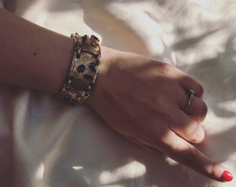Könnte beinhalten: Ein goldenes Armband mit bunten Steinen und einem strukturierten Design. Das Armband wird am Handgelenk getragen.