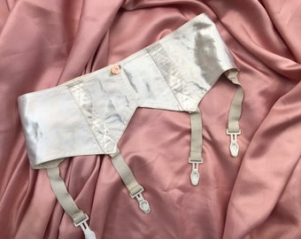 Puede incluir: Un liguero de satén blanco con tres tirantes ajustables y clips de plástico blanco. El liguero tiene un delicado adorno floral.