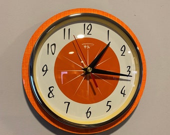 May include: Orange and white analogue clock with black hands and a gold rim. The clock face has a stylised orange slice design with a white background and black numbers.