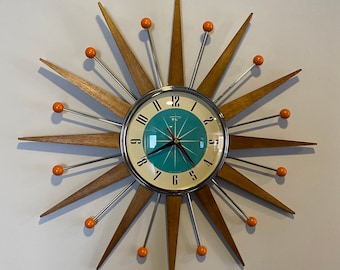 Puede incluir: Un reloj de pared de estilo moderno de mediados de siglo con un diseño de estrella. La esfera del reloj es azul turquesa con números y manecillas plateados. La estrella está hecha de madera con acentos naranjas.
