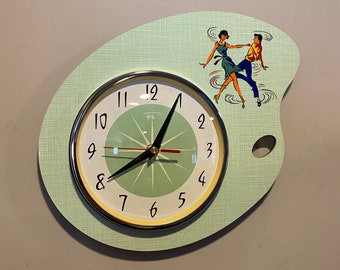 May include: A green and white clock shaped like a painter's palette with a couple dancing on the palette. The clock face has black numbers and hands.
