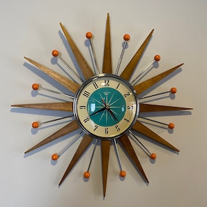 May include: A mid-century modern wall clock with a starburst design. The clock face is teal blue with silver numerals and hands. The starburst is made of wood with orange accents.