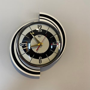 May include: A black and white clock with a chrome face and gold hands. The clock is shaped like a circle with a curved, black and white design on the side. The clock face has black numbers and a black circle in the center with the words "Made in England" printed on it.