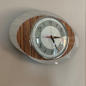 Peut inclure: Un cadran d'horloge rond et blanc avec des chiffres et des aiguilles noirs. L'horloge est encadrée d'argent et montée sur une plaque ovale en bois avec un motif rayé brun clair et gris.