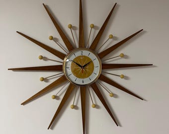 May include: A starburst wall clock featuring a central circular face with a mustard yellow and white dial, black numerals, and thin black hands. The clock has long, pointed wooden rays and shorter metal spokes tipped with small yellow spheres.