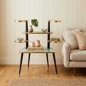 Könnte beinhalten: Ein mehrstöckiger Pflanzenständer im Mid-Century-Modern-Stil. Er hat eine hellblaue Glasplatte und Regale mit goldenen Rändern. Der Ständer wird von schwarzen Beinen mit goldenen Spitzen getragen. Auf den Regalen sind verschiedene dekorative Gegenstände wie Vasen und Bücher angeordnet.
