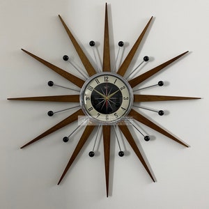 May include: A mid-century modern wall clock with a starburst design. The clock face is black with white Roman numeral hour markers and silver hands. The clock has a wooden frame with 12 wooden rays extending outward from the center. The rays are painted a dark brown color and have black ball accents at the ends.