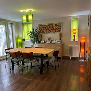 May include: A wooden dining table with six chairs in a modern dining room. The table has a light wood finish and the chairs are black metal with a light brown fabric seat. The room has a light wood floor and a large window with green blinds. There are several lamps and a large piece of art on the wall.