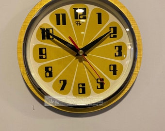 May include: A yellow and black round wall clock with a lemon design. The clock face has black numbers and hands. The clock has a silver rim and a yellow outer ring. The text on the clock reads "Royale Sterburst Clocks UK Designed by David Brege ONLY AVAILABLE TO PURCHASE FROM ROW IN THE UK".