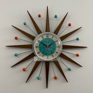 May include: A mid-century modern starburst clock with a teal face and silver hands. The clock features dark brown wooden rays, alternating with silver rods tipped with orange and teal spheres. The clock is mounted on a white wall.