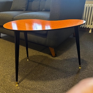Puede incluir: Mesa de centro con forma de riñón de estilo moderno de mediados de siglo. La mesa tiene una tapa naranja moteada vibrante, patas negras y puntas doradas. La mesa mide aproximadamente 122 cm de largo.