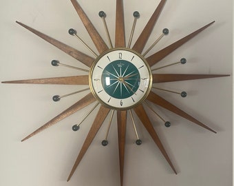 Puede incluir: Un reloj de pared de estilo moderno de mediados de siglo con una esfera verde y detalles dorados. El reloj tiene un diseño de estrella con rayos de madera y detalles negros.