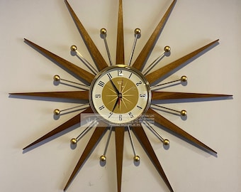 May include: A gold and brown starburst clock with a white face and black hands. The clock has a gold rim and gold accents around the numbers. The clock is mounted on a white wall.