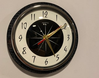 May include: A black and white clock with a black face and white numbers. The clock has a gold hour hand and a red minute hand. The clock is hanging on a white wall.