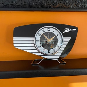 May include: A black and white clock with a silver face and black hands. The clock is shaped like a retro radio and has the word "ZENITH" printed on the side. The clock is sitting on a black shelf.